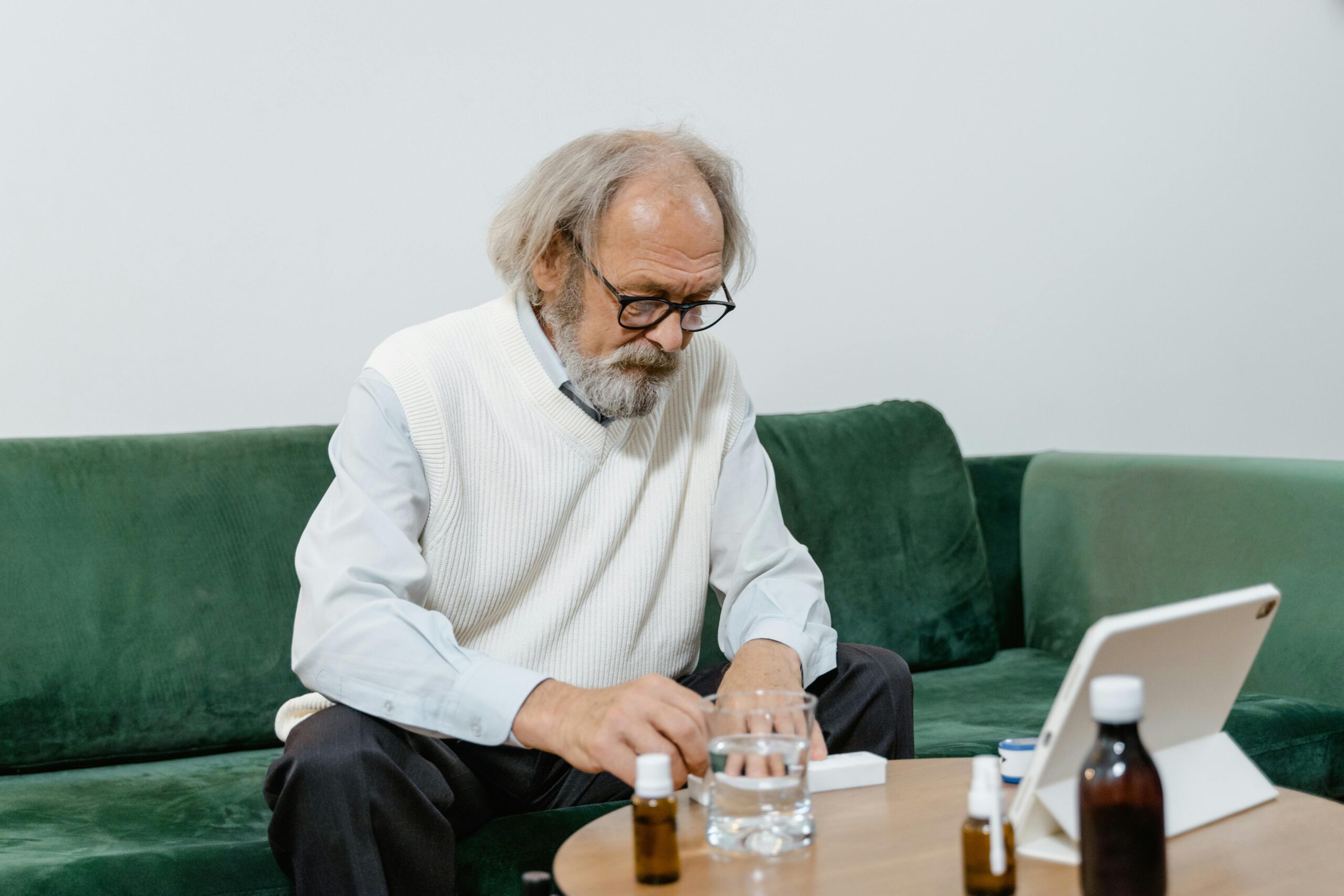 An elderly man sitting on a sofa, taking medication with water.