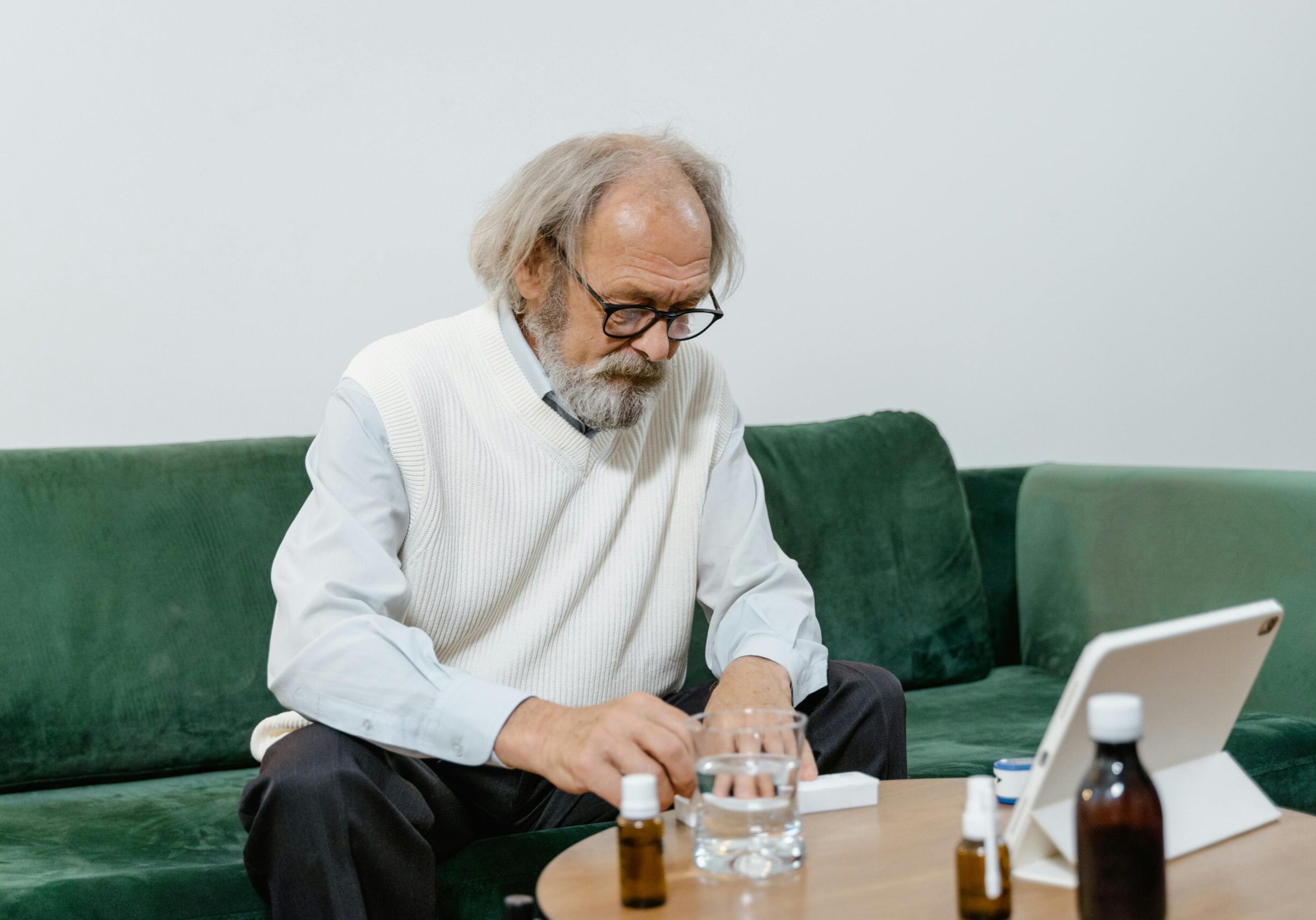 An elderly man sitting on a sofa, taking medication with water.
